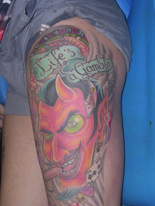 Red Ink Demon Head With LifeвЂ™s A Gamble Banner Tattoo On Left Arm